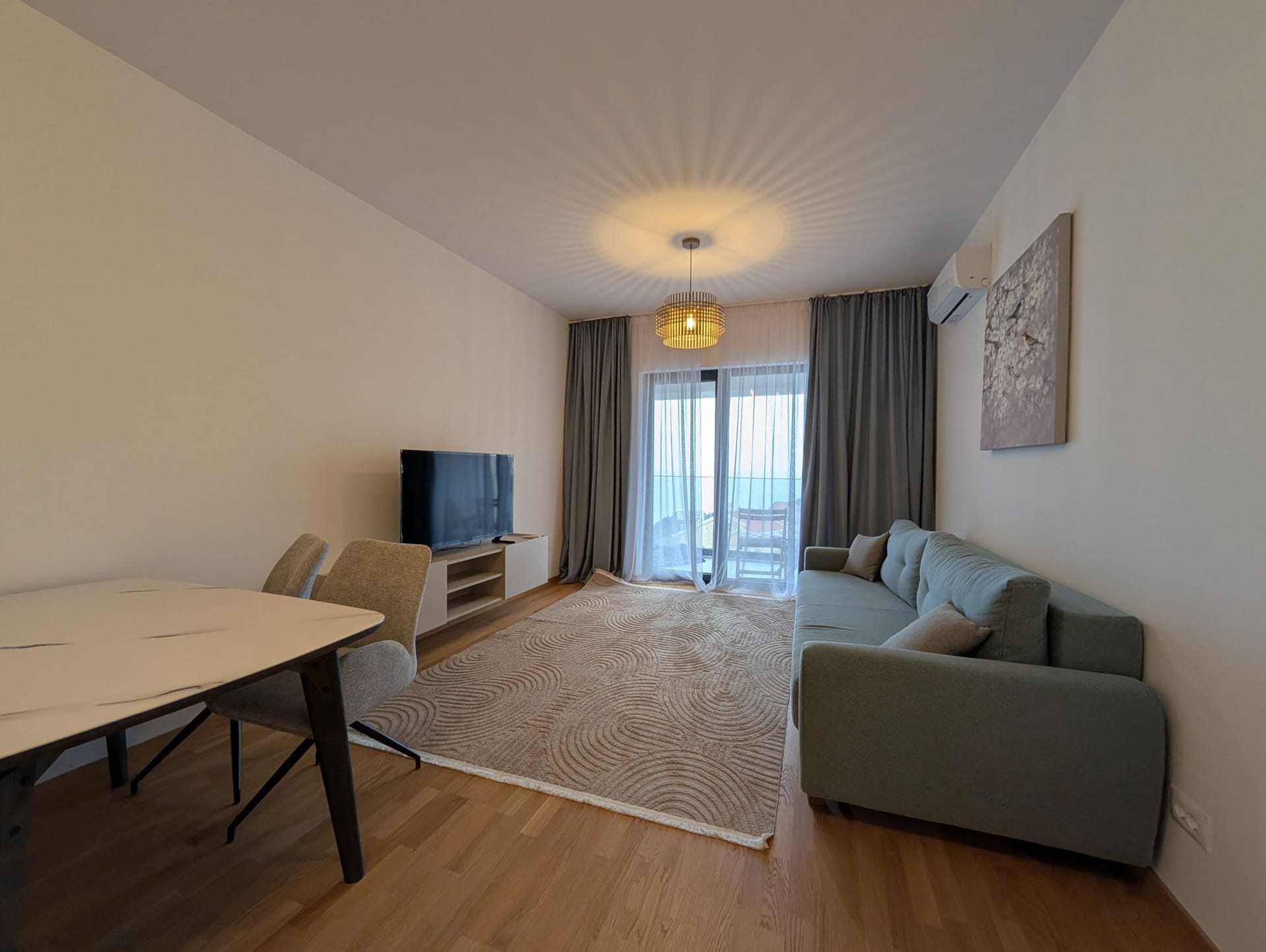 Modern living room in Bečići apartments, Montenegro: sofa, dining area, TV, and access to a terrace with a sea view.