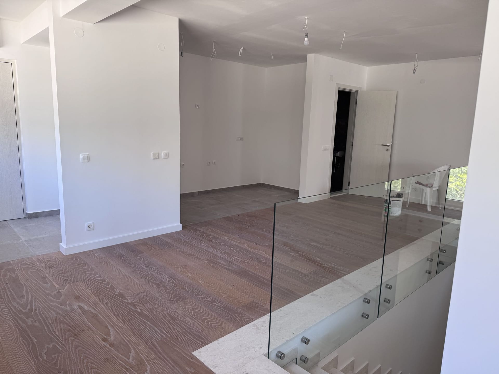 Bright living room in a new building in Kotor, Montenegro: wooden floor, glass staircase railing, and white walls.