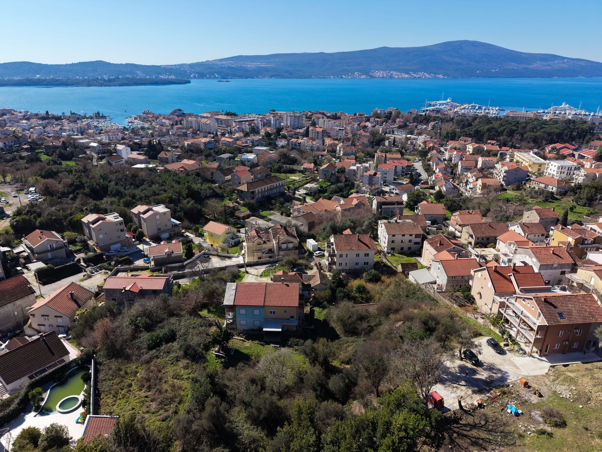 Panoramic view of Tivat, Montenegro: a plot of land with houses and a sea bay surrounded by mountains.