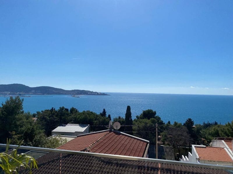 Panoramic view of the Adriatic Sea and mountains from the balcony of a house in Šušanj, Montenegro, with a terrace and tiled roofs.
