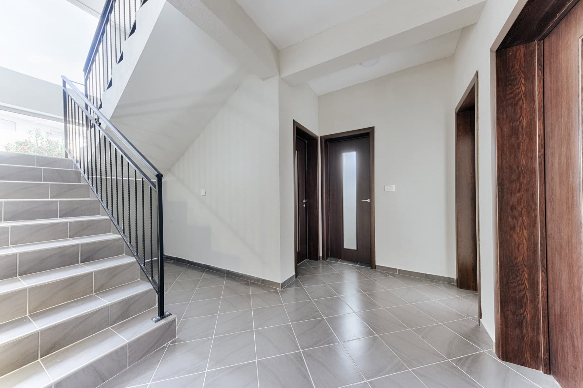 Staircase with wrought-iron railings and a bright villa hall in Utjeha, Montenegro, with gray tile finishes and dark doors.