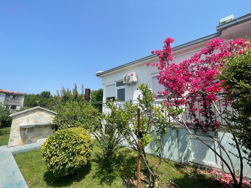 House facade in Shushan, Montenegro: blooming bougainvillea bushes, a green garden, and a stone summer kitchen in the yard.