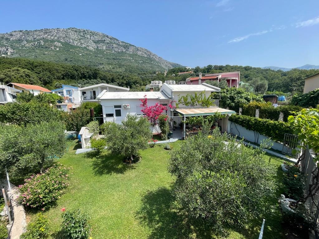 White house with a terrace and a green garden in Shushan, Montenegro, against a backdrop of scenic mountains and olive trees.