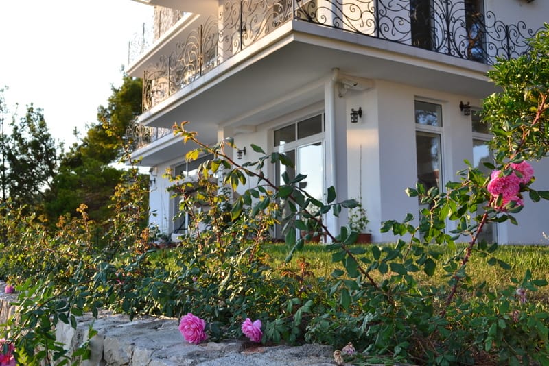 White villa with a wrought-iron balcony and a blooming garden in Shushan, Montenegro: view of the house facade and landscaped grounds.