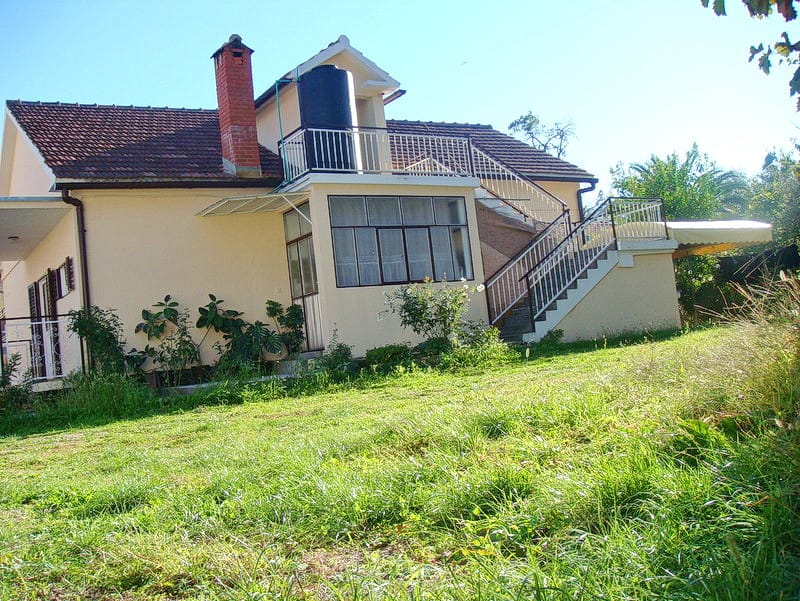 Two-story house with a terrace and exterior staircase in Baošići, Montenegro, surrounded by a green garden and plot.