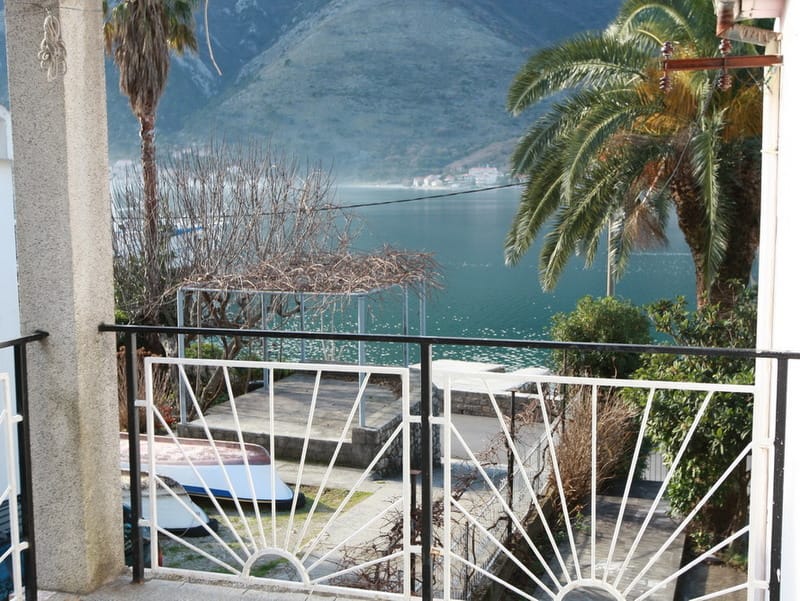 View from the balcony of a house in Dobrota, Montenegro: a panorama of the Bay of Kotor, mountains, a palm tree, and boats on the shore.
