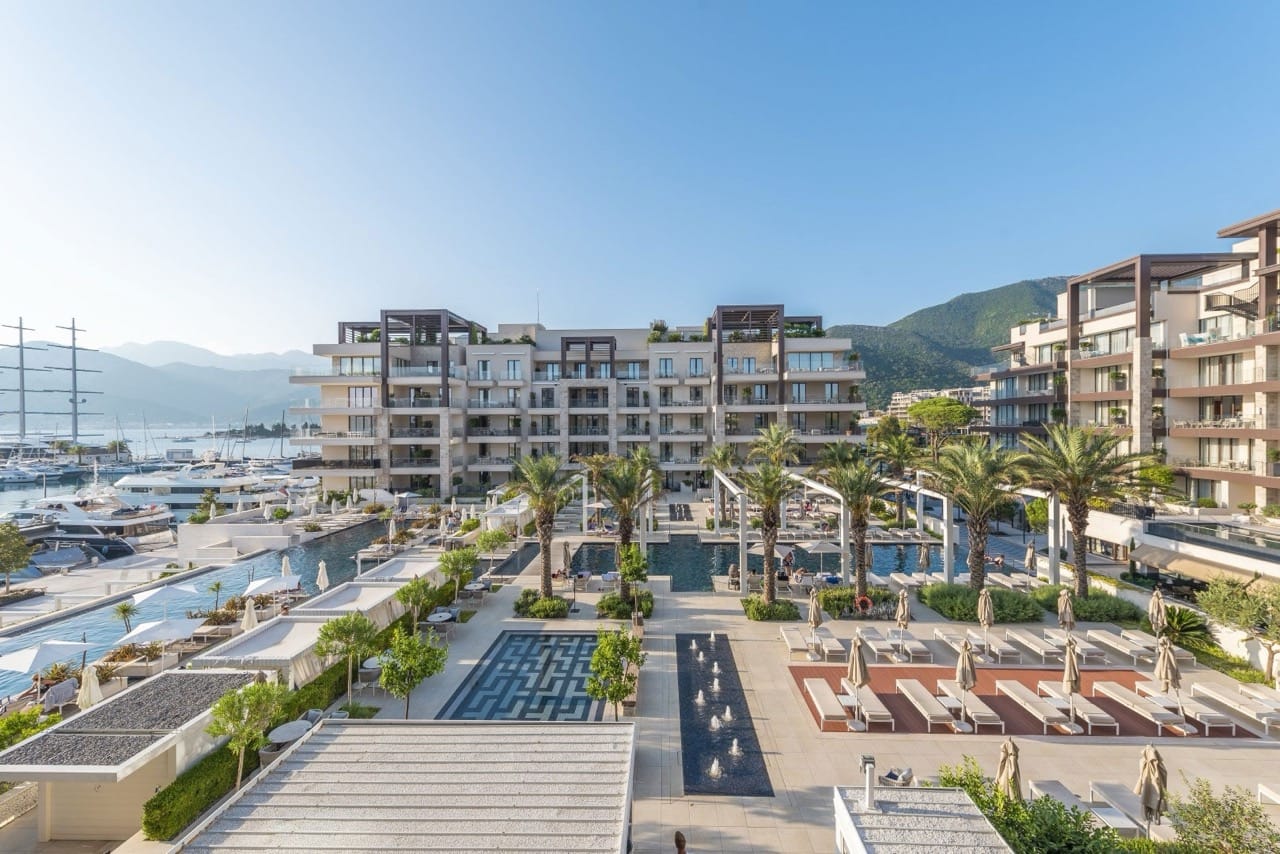 View of the pool and luxury residential complex in Porto Montenegro, Tivat, Montenegro, with a yacht marina and mountains in the background.