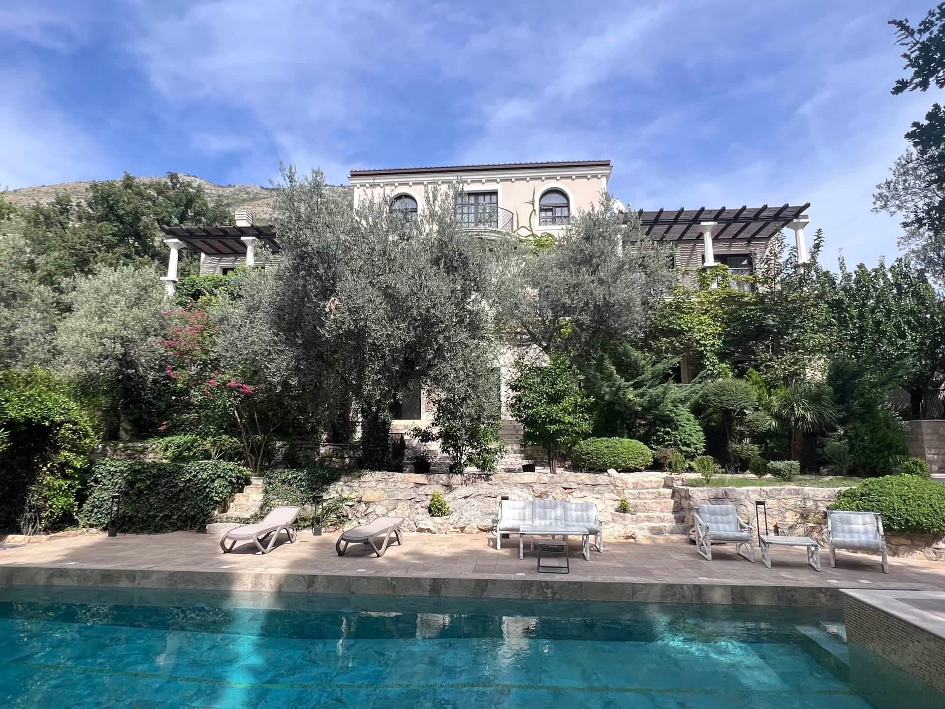 Вилла в Режевичи, Черногория: бассейн, терраса с шезлонгами и сад на фоне каменного фасада элитного дома.