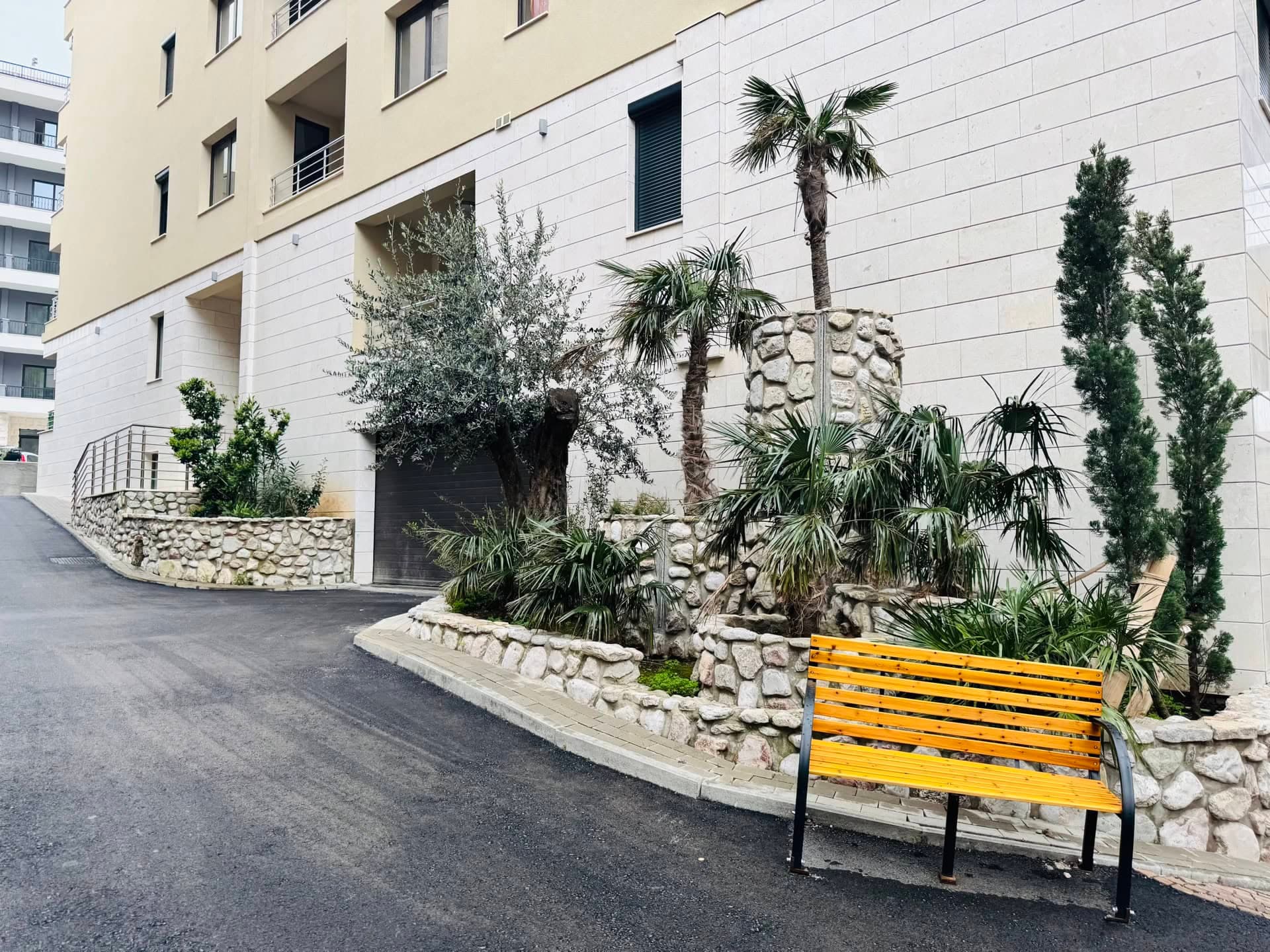 Благоустроенная территория жилого комплекса в Бечичи, Черногория: каменная отделка, пальмы и скамейка у дома.