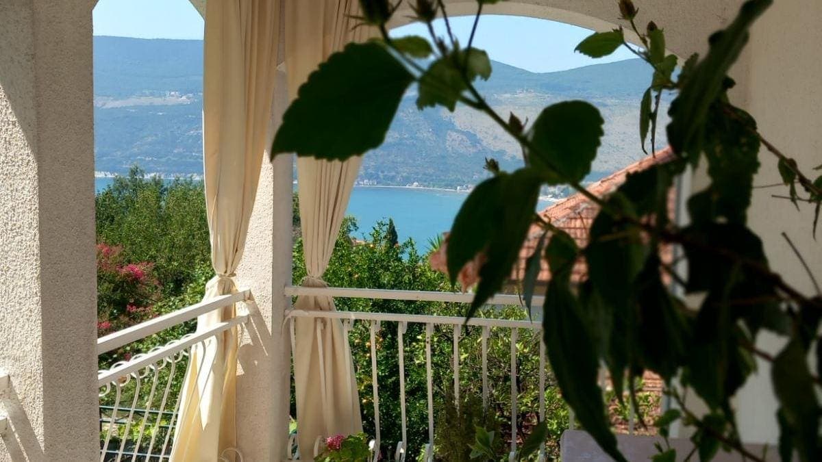 Вид на море и горы с террасы дома в Игало, Черногория: белая балюстрада, светлые шторы и зеленые растения.