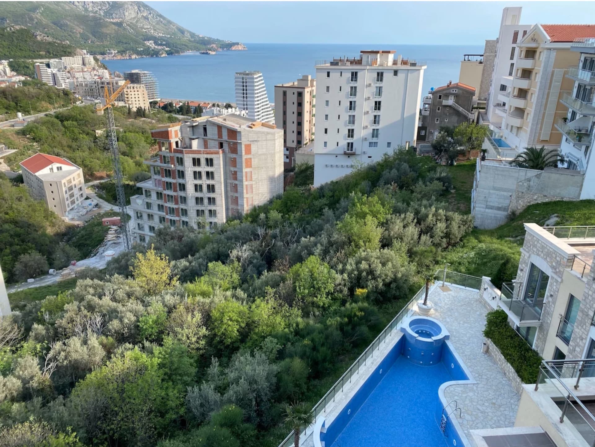 Вид с террасы квартиры в Бечичи, Черногория: бассейн, зеленый склон, городская застройка и панорама Адриатического моря.