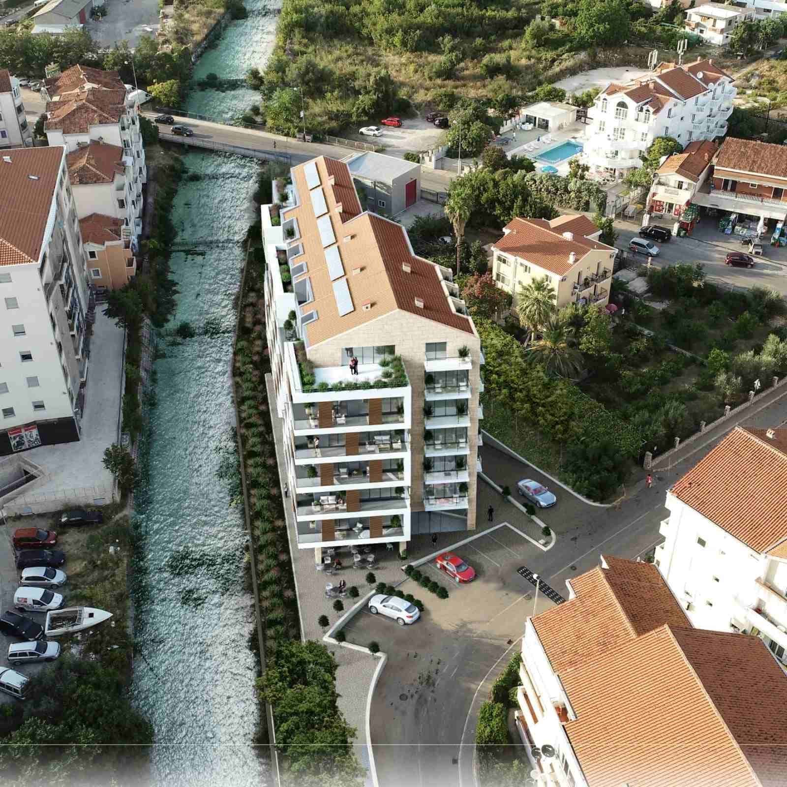 Вид с воздуха на жилой комплекс в Будве, Черногория: современное здание с террасами у реки и парковкой.