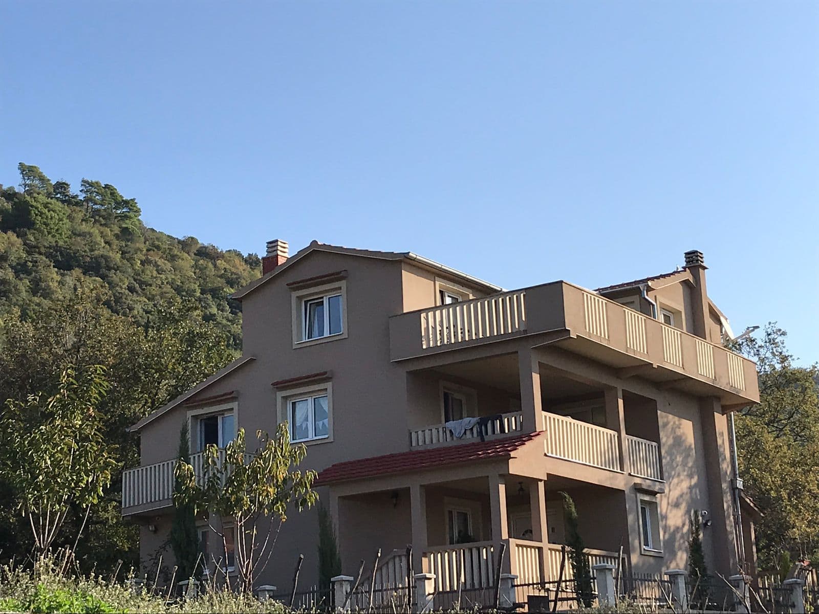 Трехэтажный дом с террасами на полуострове Луштица, Черногория, на фоне зеленого холма и ясного неба.