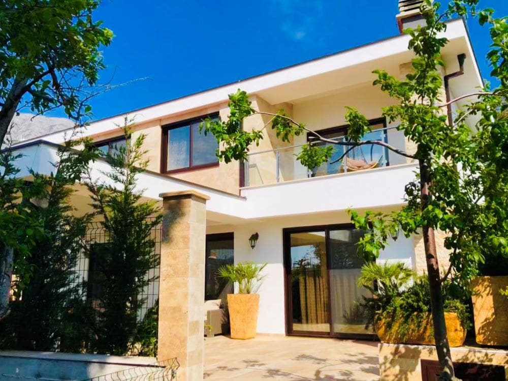 Двухэтажная вилла с террасой и садом в Добрых Водах, Черногория: современный фасад, панорамные окна и зеленый участок.