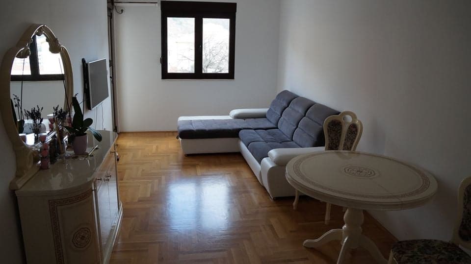 Гостиная в квартире в Будве, Черногория: серый угловой диван, обеденный стол, комод с зеркалом и паркетный пол.