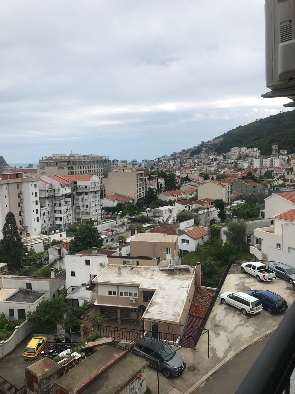 Вид с балкона квартиры в Будве, Черногория: панорама города, жилые дома, горы и парковка у здания.