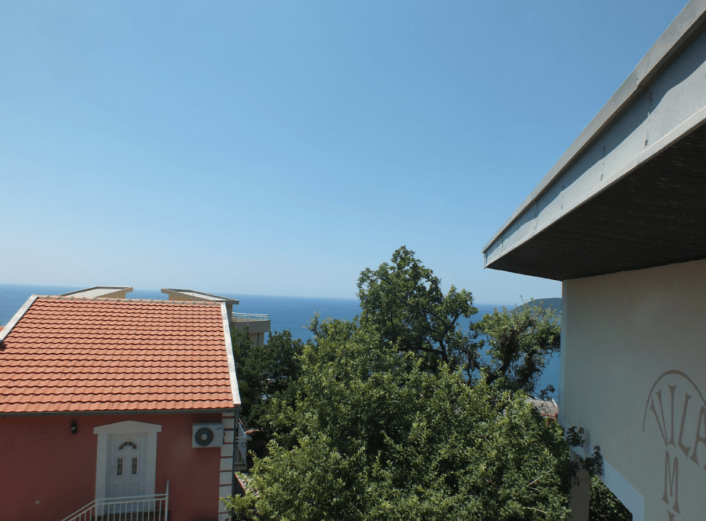 Вид на море и крыши домов с террасы дома в городе Бар, Черногория, в окружении зеленых деревьев и ясного неба.