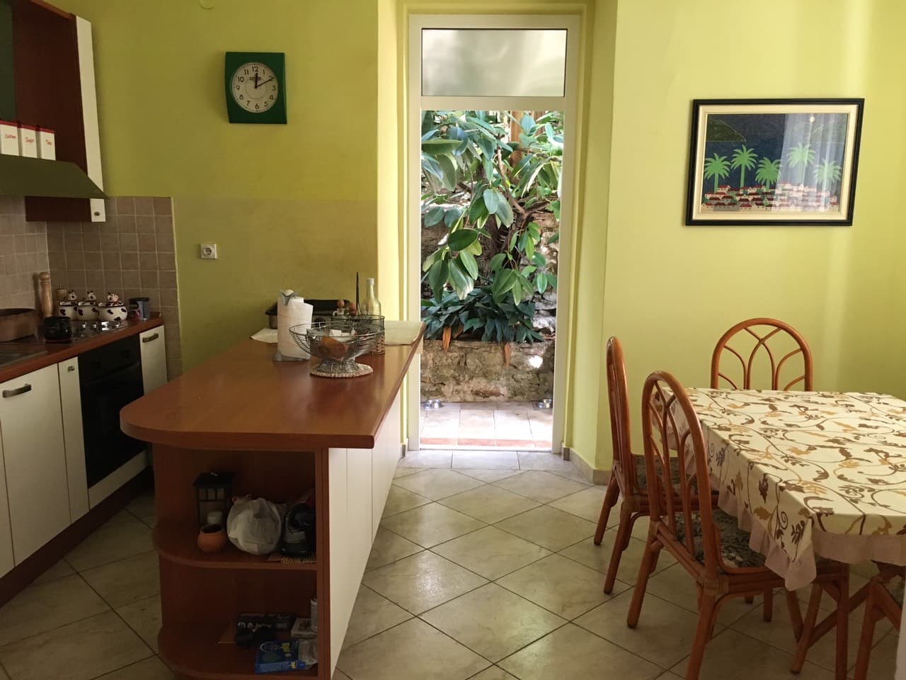 Кухня с барной стойкой и обеденной зоной в доме в Герцег-Нови, Черногория, с выходом в зеленый сад через стеклянную дверь.