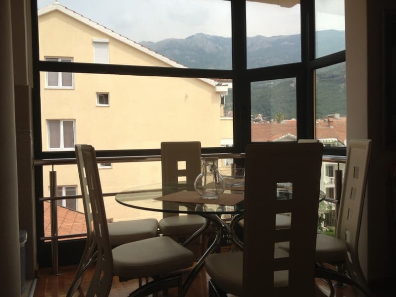 Обеденная зона в квартире в Будве, Черногория: стеклянный стол, стулья и панорамный вид на горы и город.