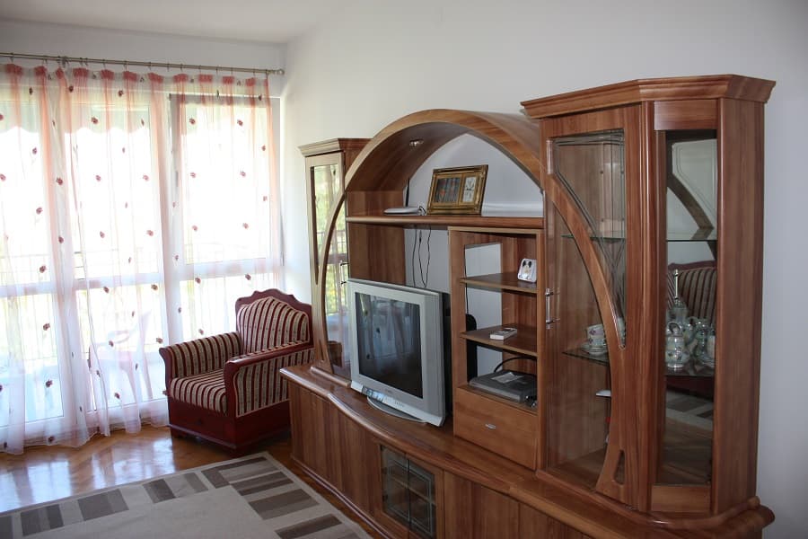 Гостиная в квартире в Будве, Черногория: деревянная стенка, телевизор, полосатое кресло и выход на балкон с тюлем.