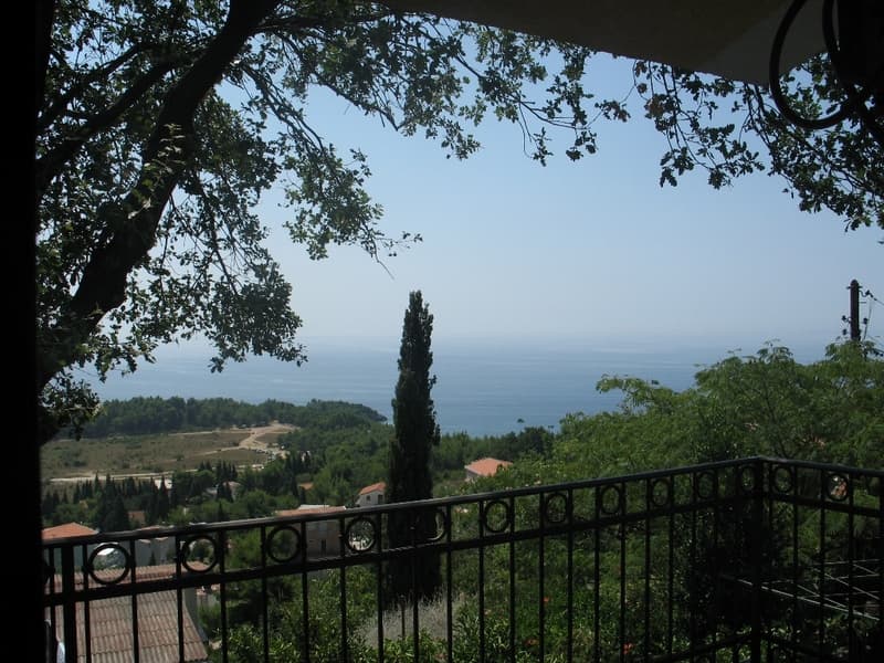 Вид на море и зеленые холмы с террасы дома в Сутоморе, Черногория, с кованым ограждением и кронами деревьев.