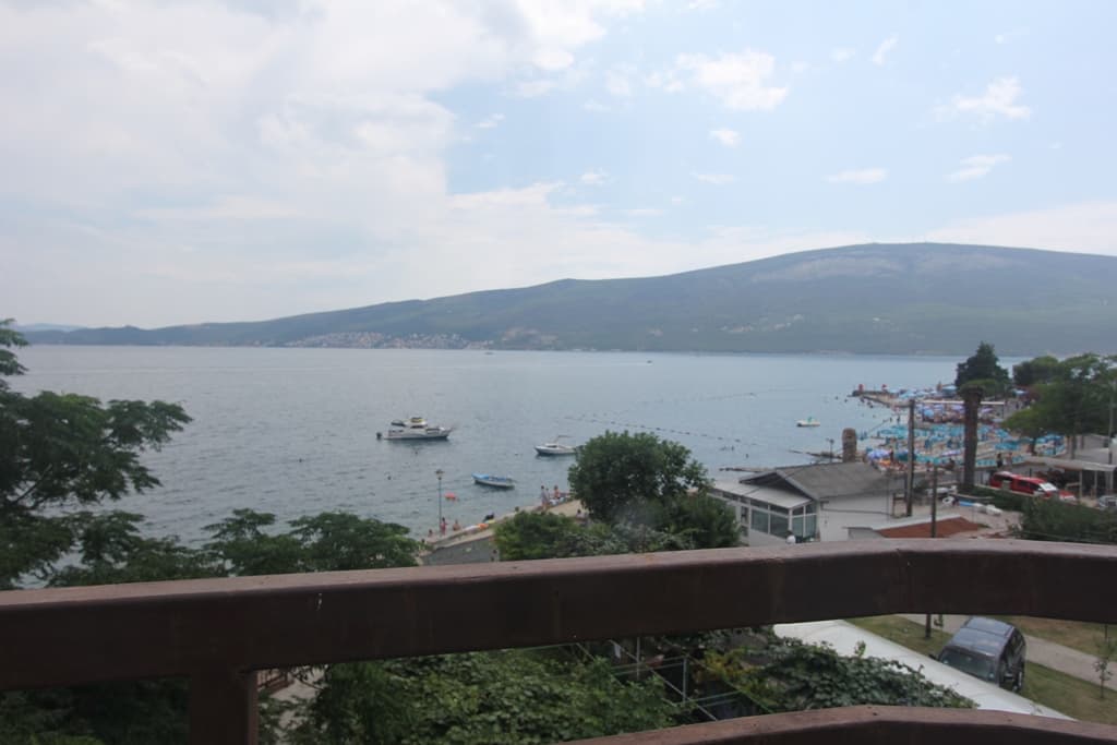 Панорамный вид на море и пляж с балкона мини-отеля в Баошичи, Черногория: горы, яхты и прибрежная инфраструктура.