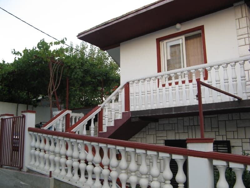 Двухэтажный дом в Шушани, Черногория: фасад с каменной отделкой, белой балюстрадой и террасой с виноградной лозой.