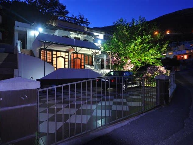 Вечерний вид на дом в Петроваце, Черногория: фасад с подсветкой, терраса, парковка и ухоженный сад у входа.