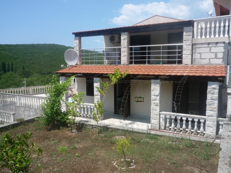 Двухэтажная вилла в Бигово, Черногория: каменная отделка, террасы с балюстрадами, сад и вид на зеленые холмы.