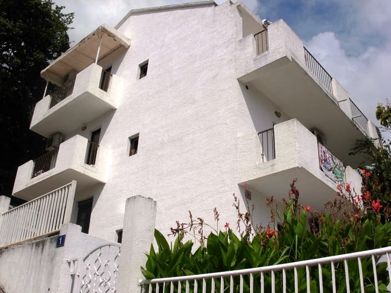 Трехэтажный белый дом с балконами в Петроваце, Черногория: фасад здания с ограждением и зелеными насаждениями.