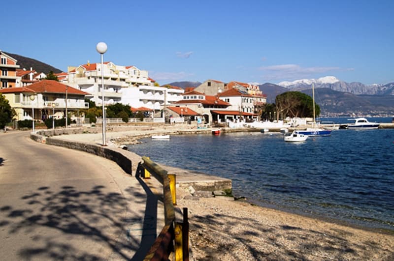 Вид на набережную и мини-отель в Дженовичи, Черногория: прибрежная дорога, пляж, яхты и заснеженные горы на фоне.