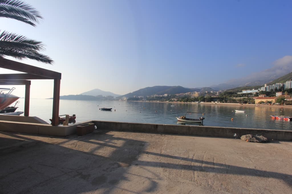 Вид на море и пляж с террасы коммерческого помещения в Рафаиловичи, Черногория: причал с лодками и горы на горизонте.