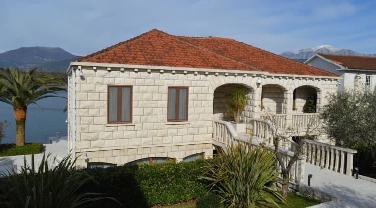 Каменная вилла в Тивате, Черногория: фасад из светлого камня, терраса с балюстрадой и вид на море и горы.