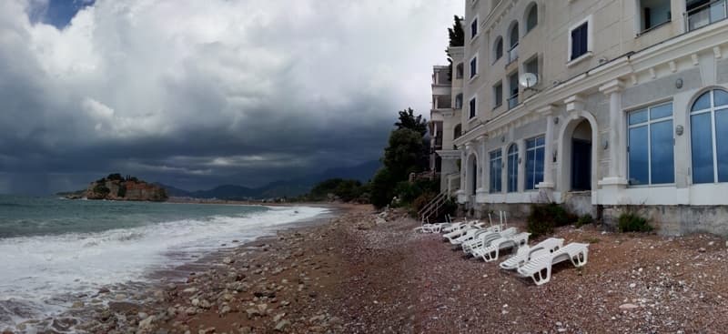 Ресторан у моря в Святом Стефане, Черногория: вид на пляж, шезлонги и остров Свети-Стефан на фоне облачного неба.