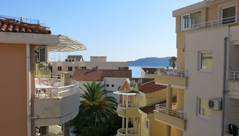 Вид с балкона мини-отеля в Рафаиловичи, Черногория: городская застройка, пальмы и морской горизонт вдали.