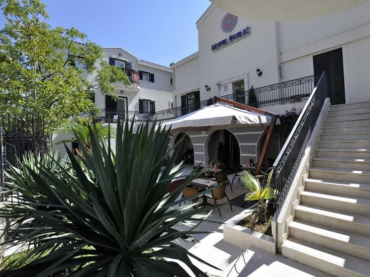 Фасад отеля Durc в Петроваце, Черногория: уютная терраса с зонтами, уличная мебель и каменная лестница в окружении зелени.