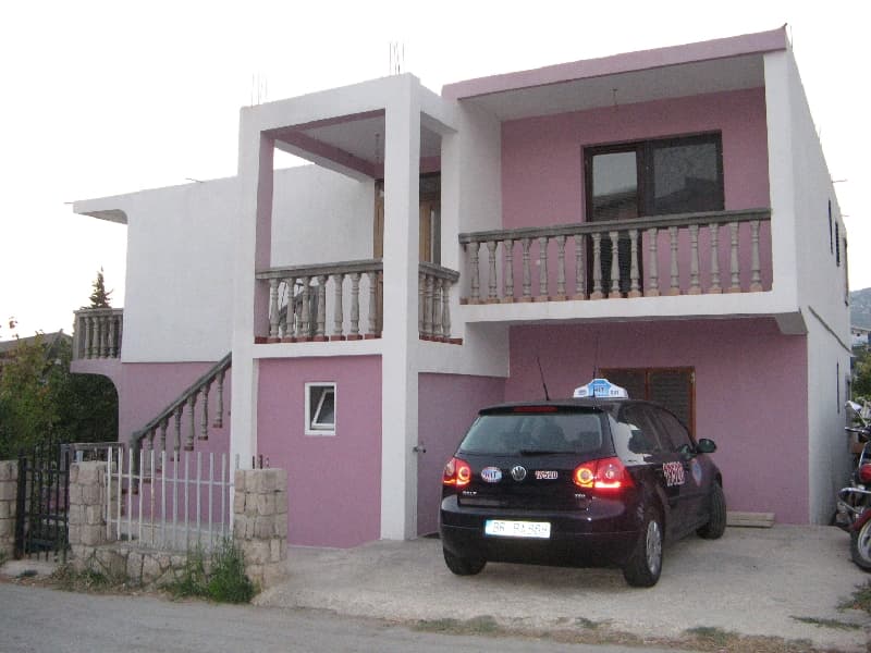 Двухэтажный дом с розовым фасадом и террасами в Шушани, Черногория, с парковкой для автомобиля перед входом.