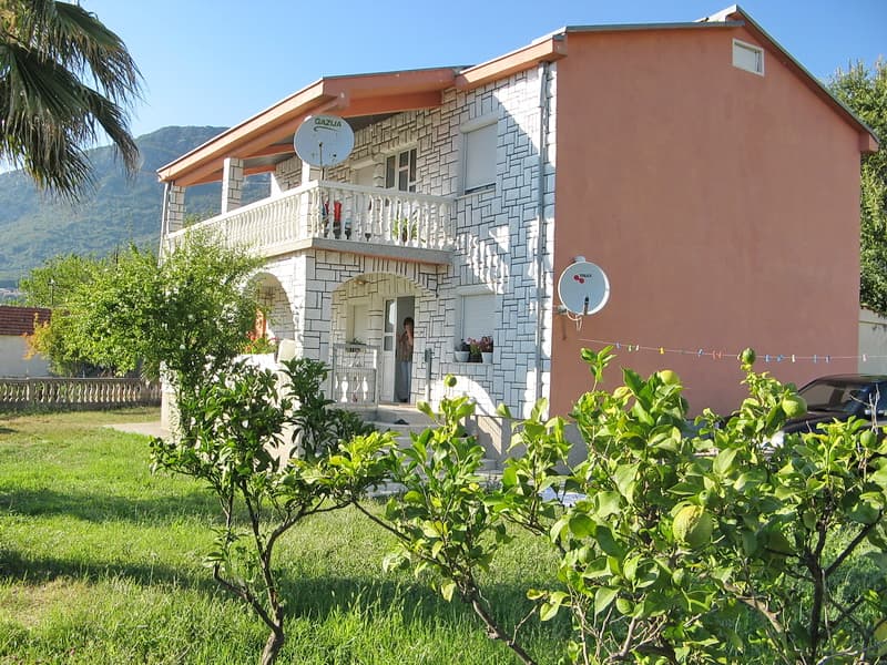 Двухэтажный дом с каменной отделкой и балконом в городе Бар, Черногория, на фоне гор и зеленого сада.