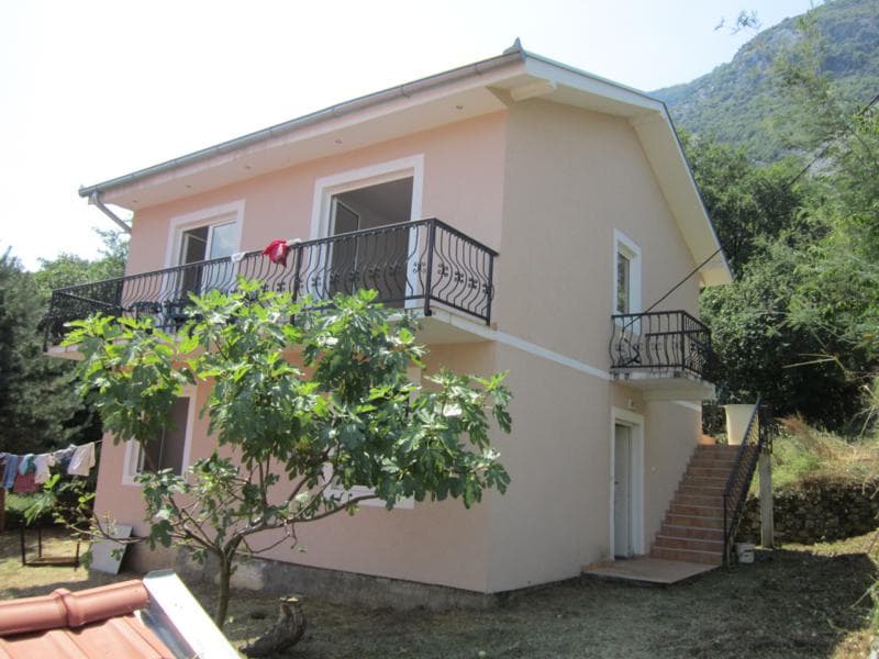 Двухэтажный дом с балконами и коваными перилами в Которе, Черногория, окруженный садом и горным пейзажем.