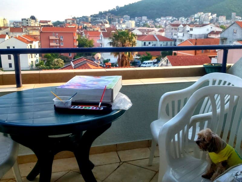 Вид с террасы квартиры в Будве, Черногория: пластиковая мебель, принадлежности для рисования и собака на фоне города.