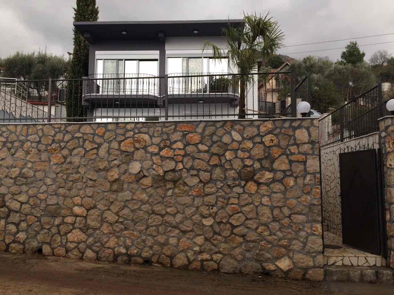 Современный дом в Шушани, Черногория: фасад с панорамными окнами, балконом и высокой каменной оградой участка.