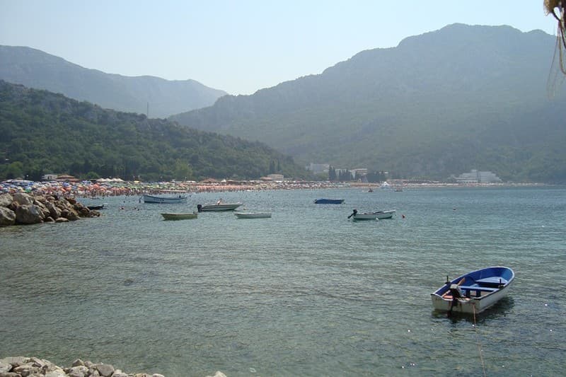 Вид на море и пляж отеля в Чани, Черногория: лодки на воде, горный пейзаж и побережье с отдыхающими в солнечный день.