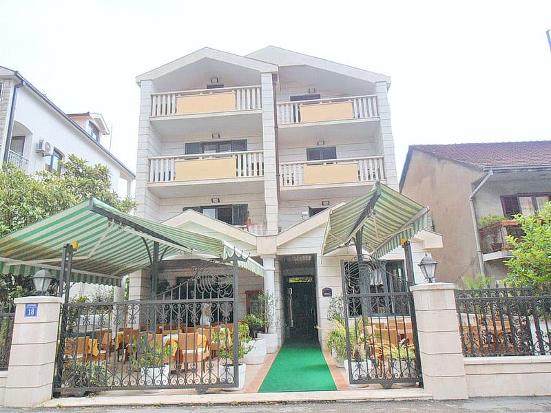 Трехэтажный отель в Тивате, Черногория, с террасой, навесами и кованым забором на главном фасаде здания.