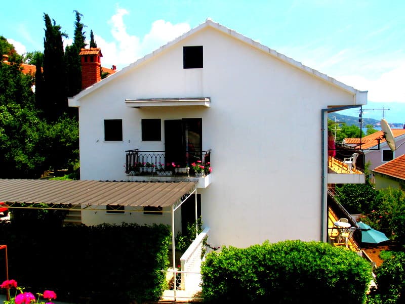 Белый двухэтажный дом с балконами и террасой в Герцег-Нови, Черногория, окруженный зеленью и деревьями.