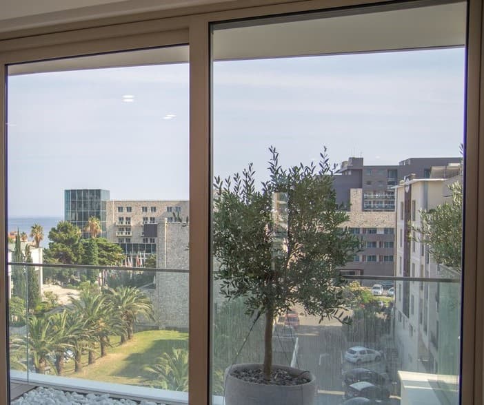 Вид из окна квартиры в Будве, Черногория: панорамное остекление, оливковое дерево на балконе и морской пейзаж.