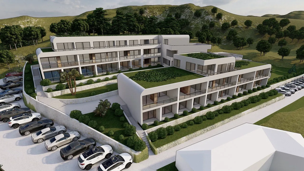 Aerial view of a modern white apartment complex with green roofs, balconies, and a parking lot against hills.