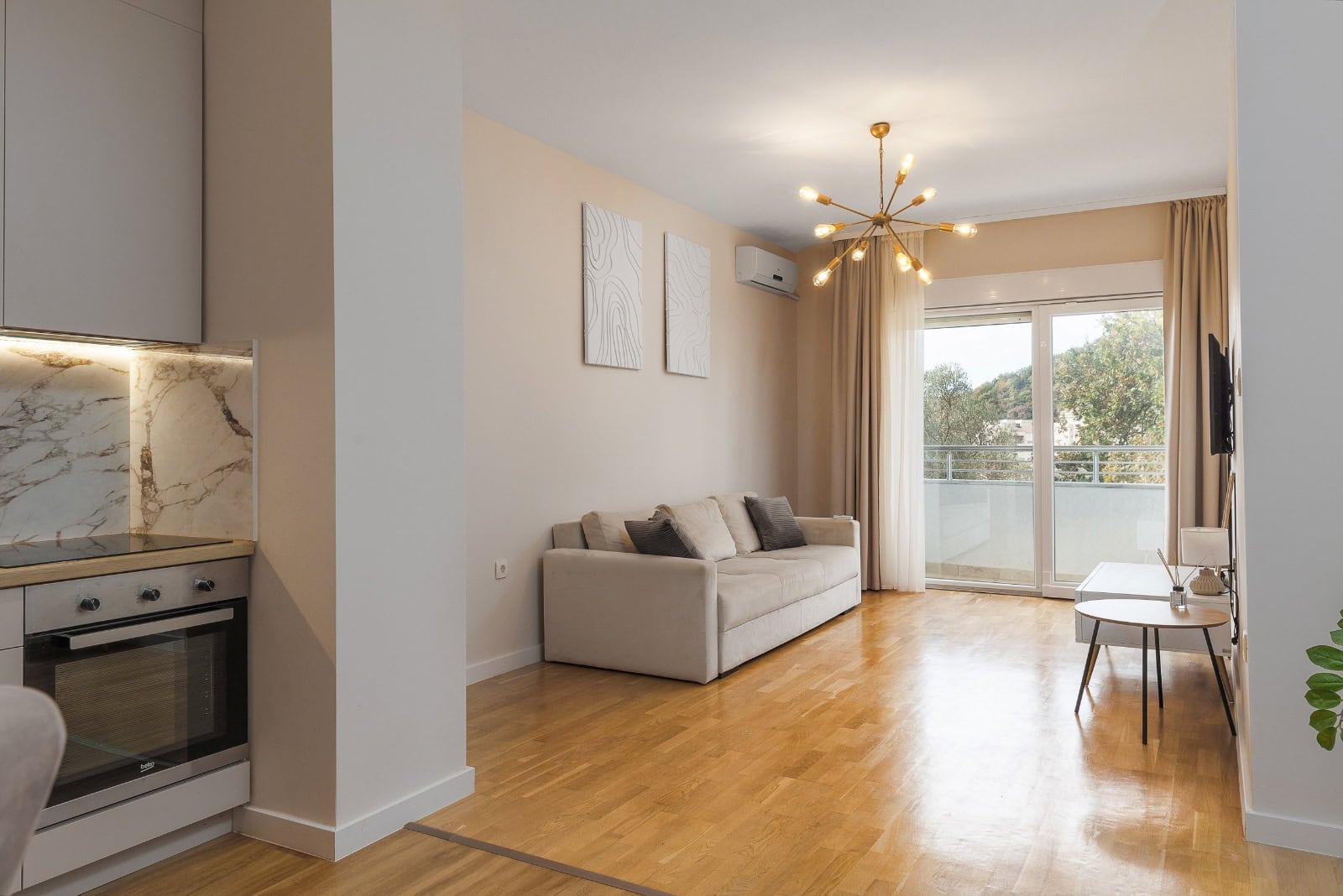 Modern living room with kitchen in an apartment in Bečići, Montenegro: light furniture, parquet flooring, and access to a balcony with a view.