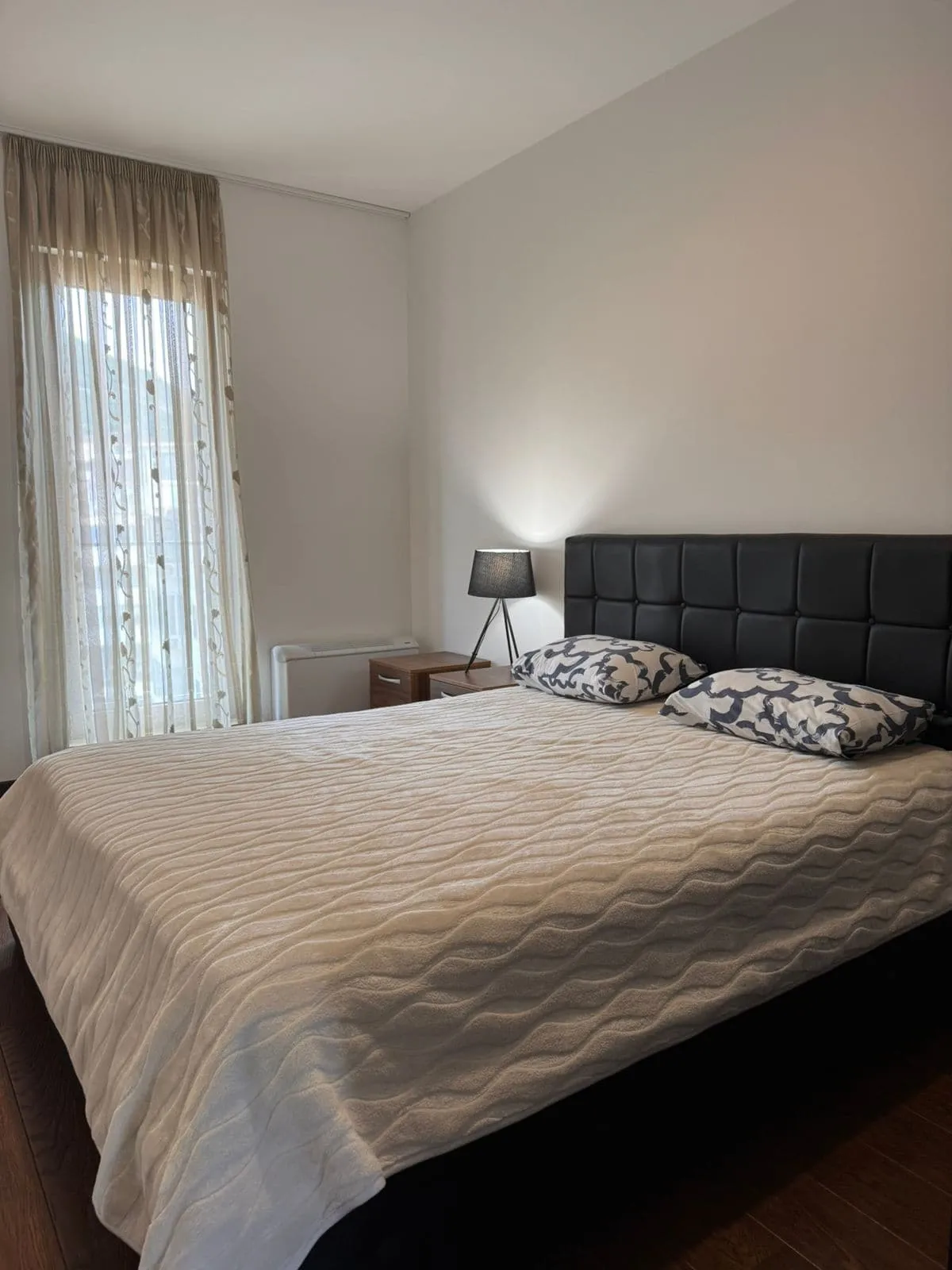 Bedroom featuring a double bed with a black upholstered headboard, nightstand, lamp, and sheer curtains.
