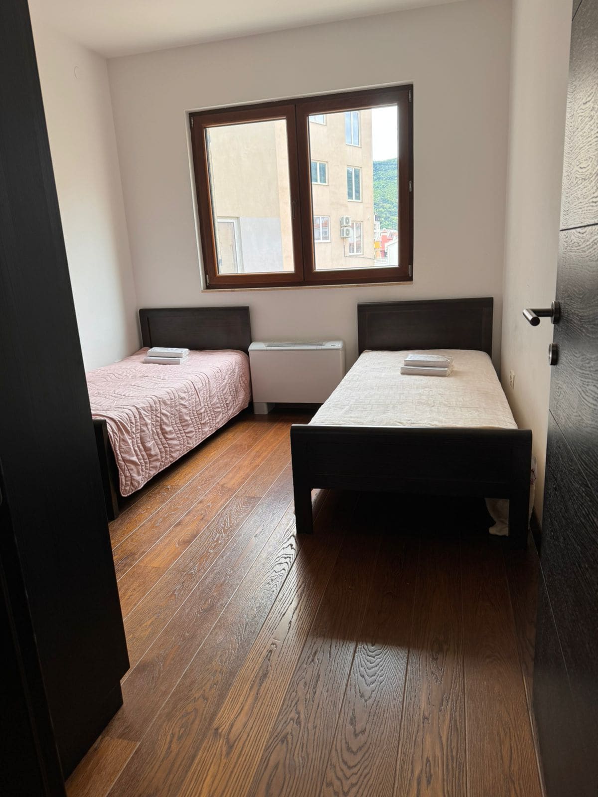 Bedroom with two beds, dark furniture and parquet flooring in an apartment in Budva, Montenegro. City view from the window.