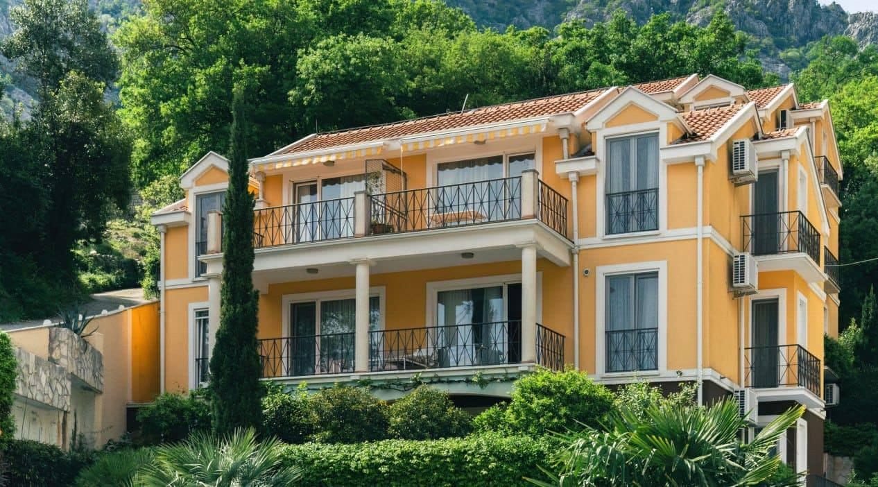 Three-story yellow house with balconies and a tiled roof in Orahovac, Montenegro, surrounded by green trees.